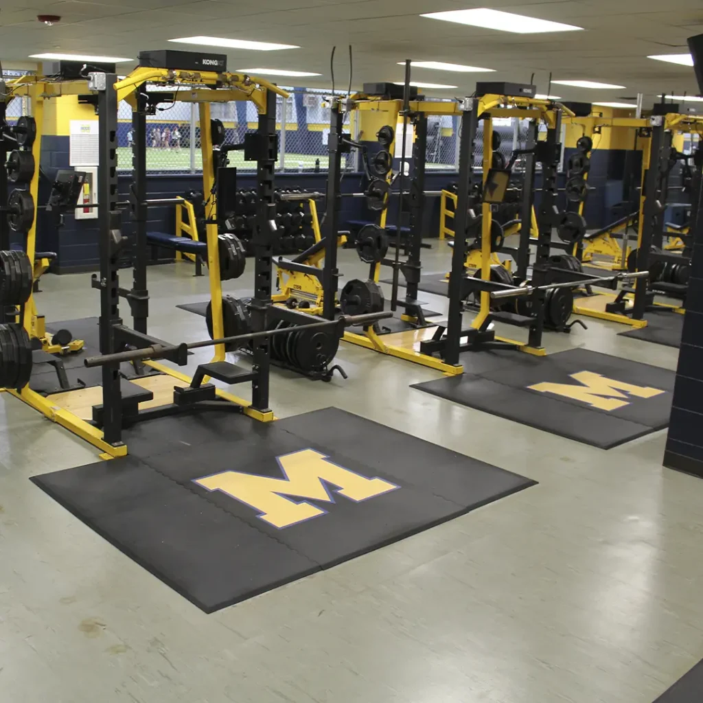Wide view of a school weight room featuring multiple rubber weightlifting platforms, several racks, and engineered “M” floor logos.