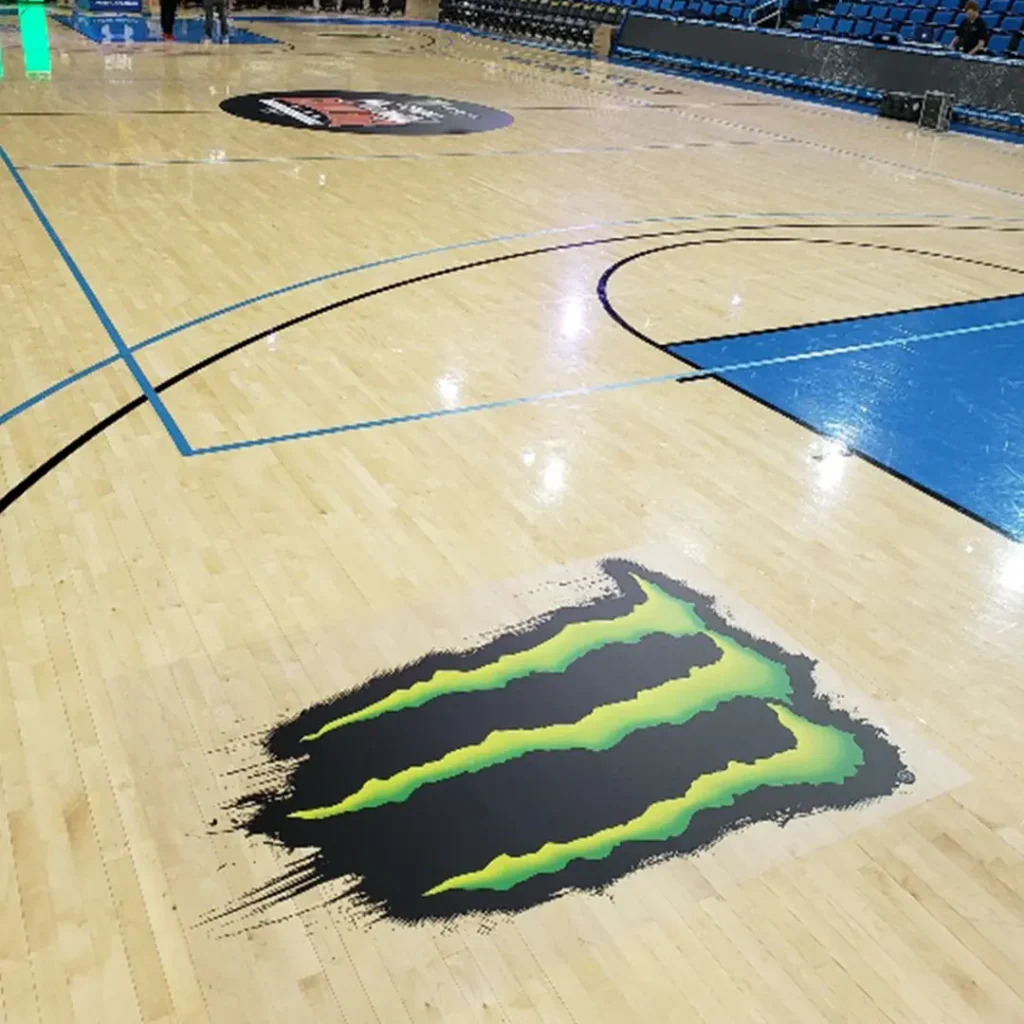Two engineered floor logos installed on a basketball court hardwood for a Monster Energy event, featuring the Monster Energy “M” logo in the foreground.