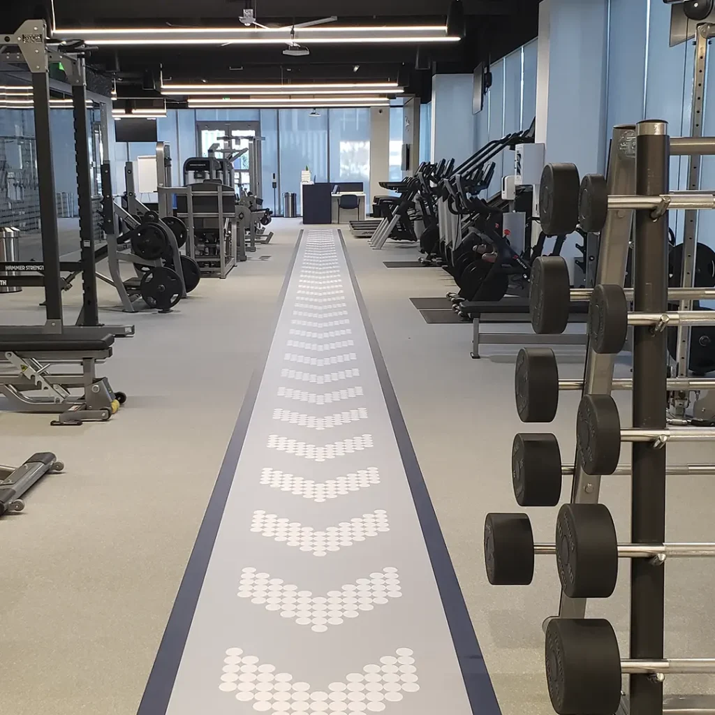 Wide view of engineered chevron traffic-flow floor graphics installed inside a fitness facility, showing directional pathways and overall space layout.