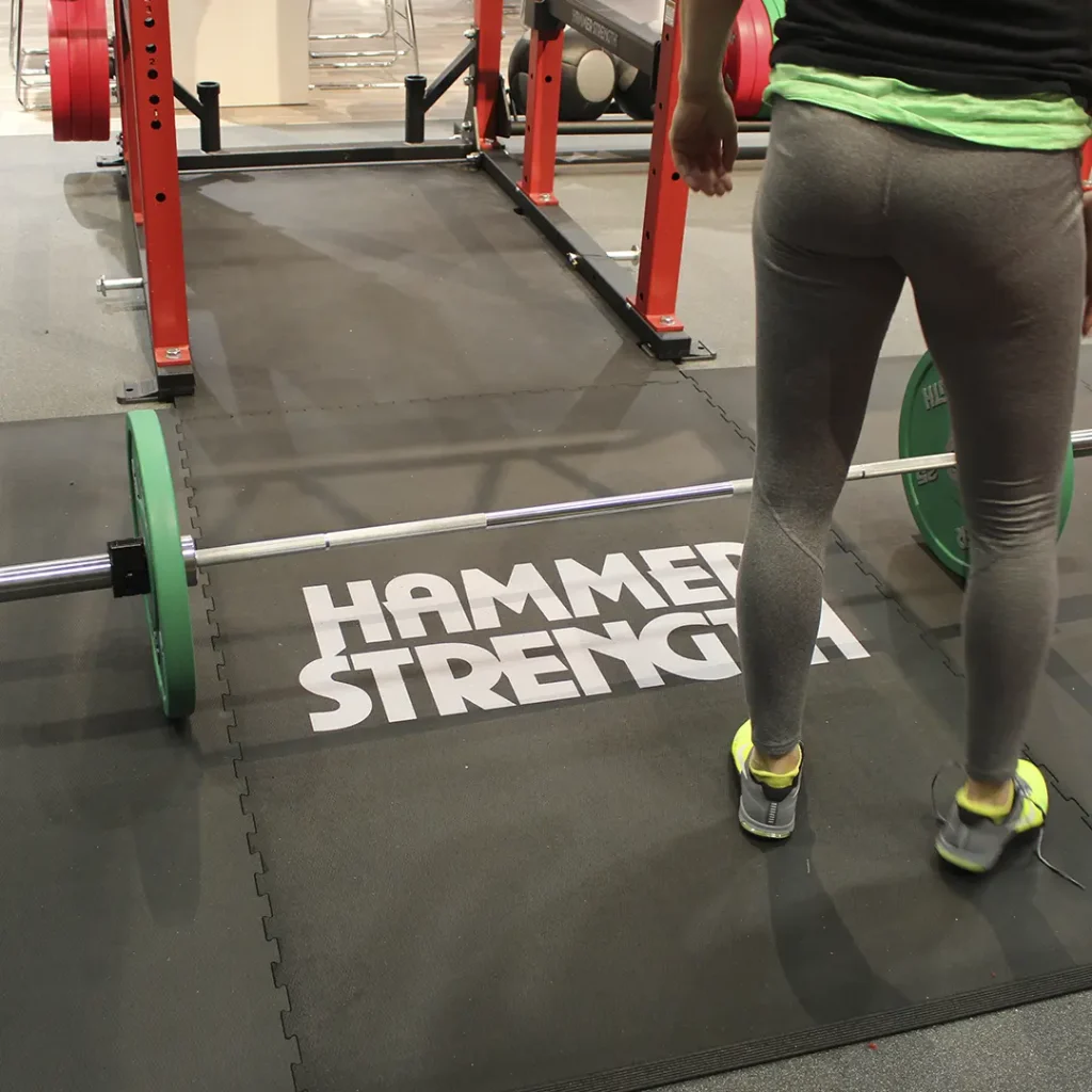 White “Hammer Strength” engineered floor logo installed on an American Platforms weightlifting platform, with a red rack, a green weight plate, and an athlete’s legs visible.
