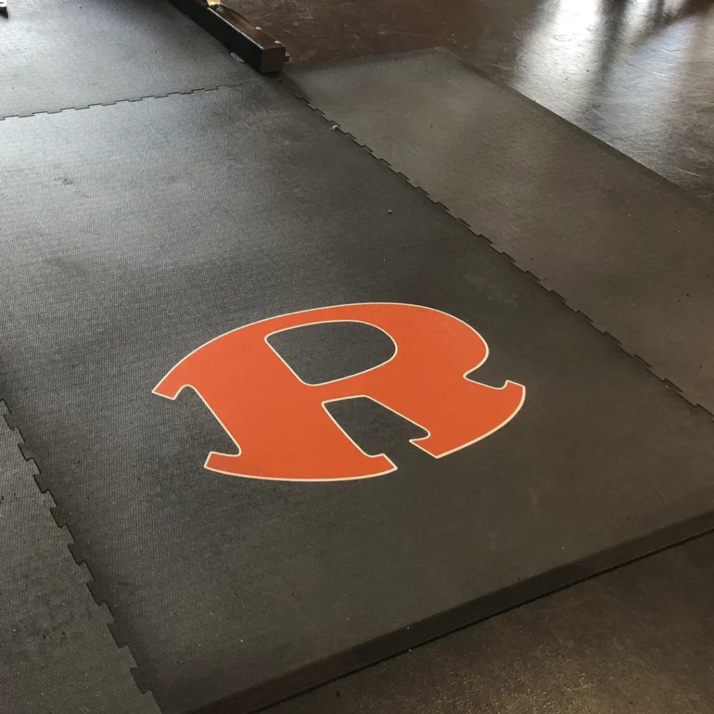 Close-up of an orange “R” engineered floor logo installed on one of our weightlifting rubber platforms, with part of a rack foot visible at the top of the image.