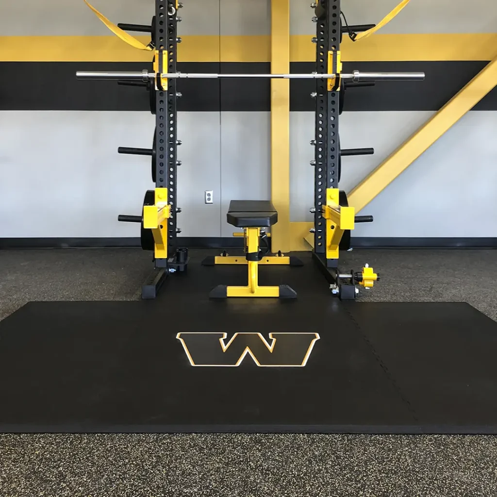 Engineered “W” floor logo installed on an American Platforms rubber weightlifting platform, with a black rack featuring yellow J-hooks and yellow safety bars.
