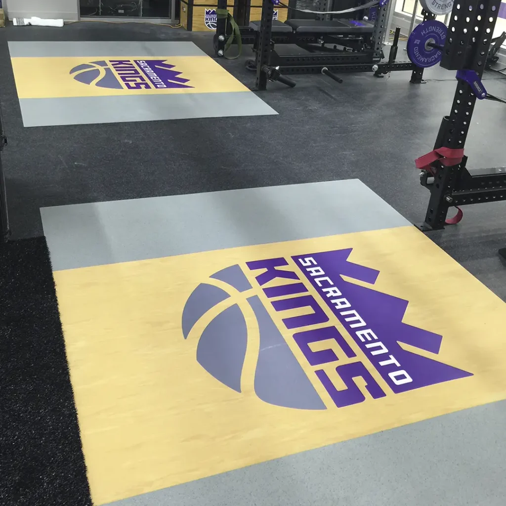 Two Sacramento Kings engineered floor logos installed on rubber and vinyl inlaid platforms, with black turf and the lower sections of two racks visible.
