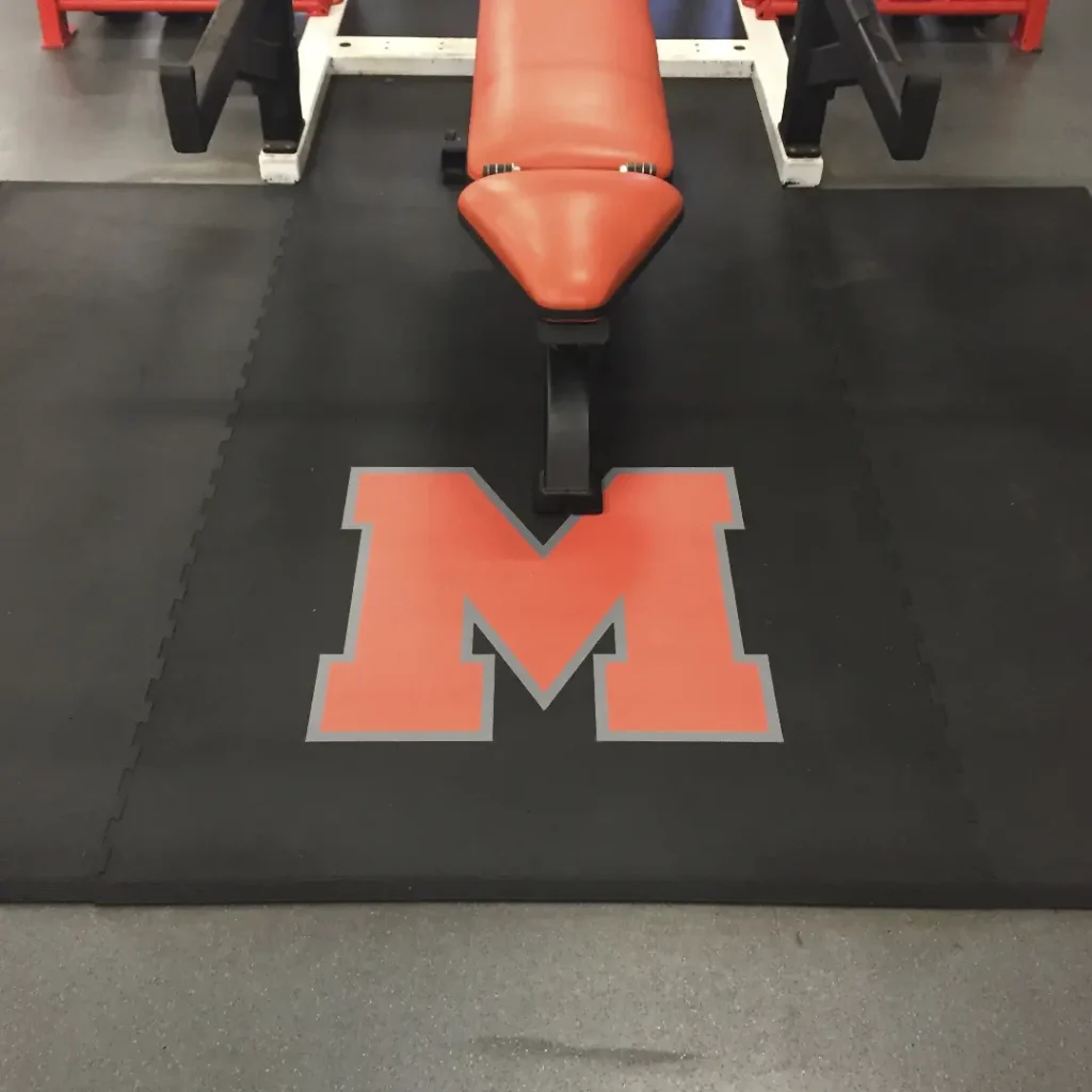 Close-up of an orange “M” engineered floor logo on a rubber weightlifting platform, with part of the rack base and a half-visible orange bench.