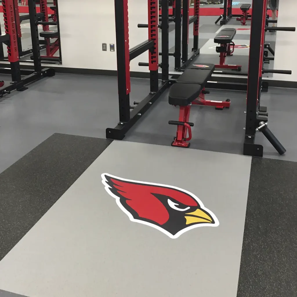 Close-up view of a red bird engineered floor graphic with a yellow beak and black face, installed on a rubber inlaid platform with part of a rack and bench visible.