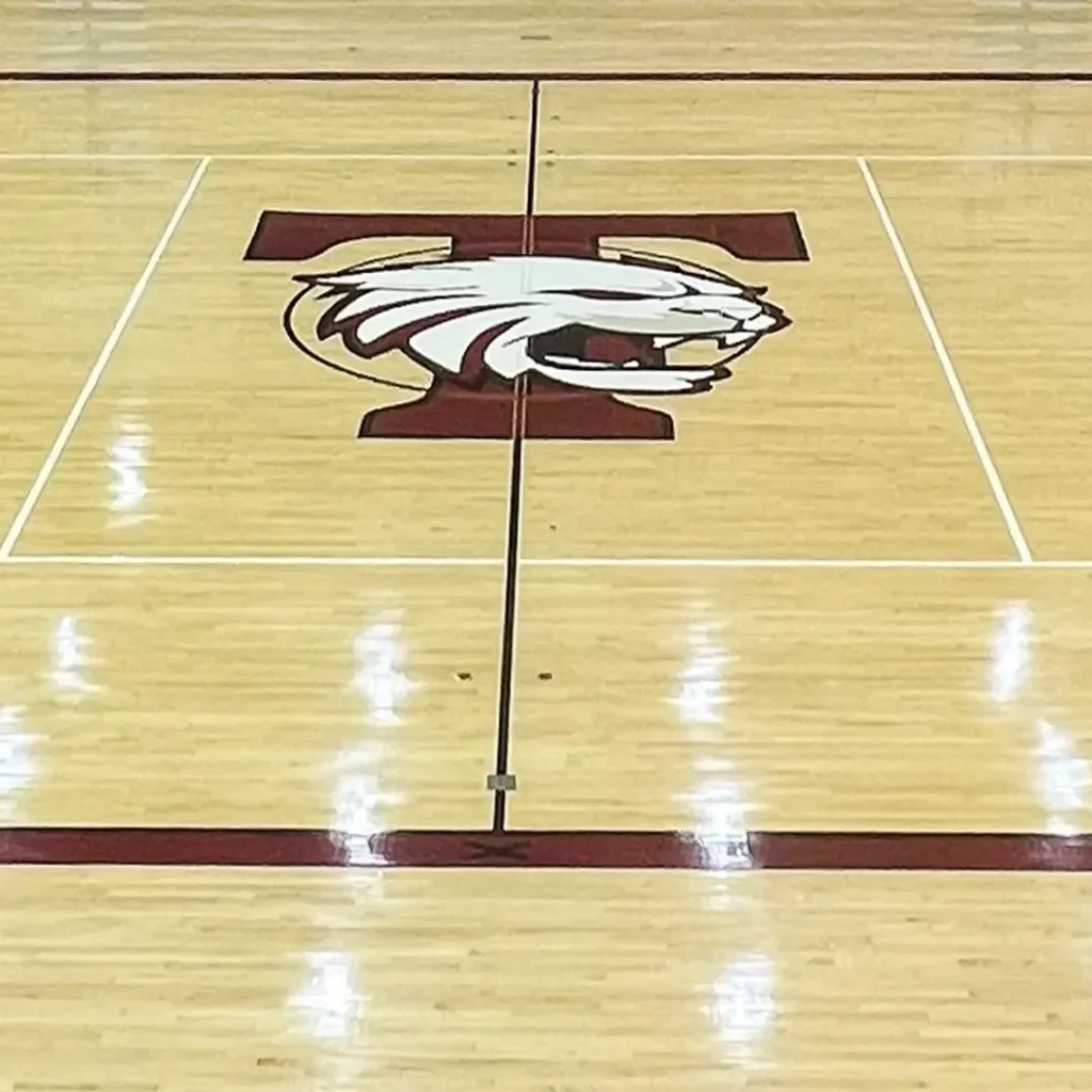 Hardwood basketball court featuring an engineered floor logo of a “T” with a wildcat graphic, with court lines partially visible.