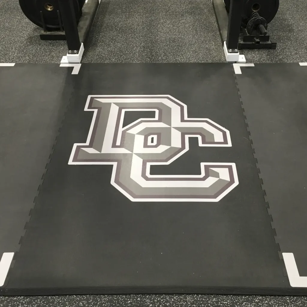Close-up view of a “DC” engineered floor logo installed on a rubber weightlifting platform, with drop zone markings visible on the sides.