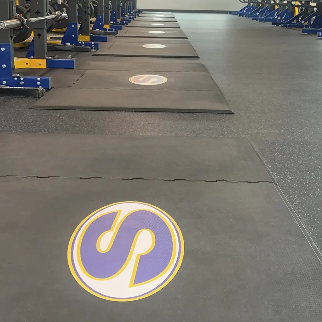 School weight room featuring engineered “S” floor logos installed on rubber weightlifting platforms, with racks visible in the training area.