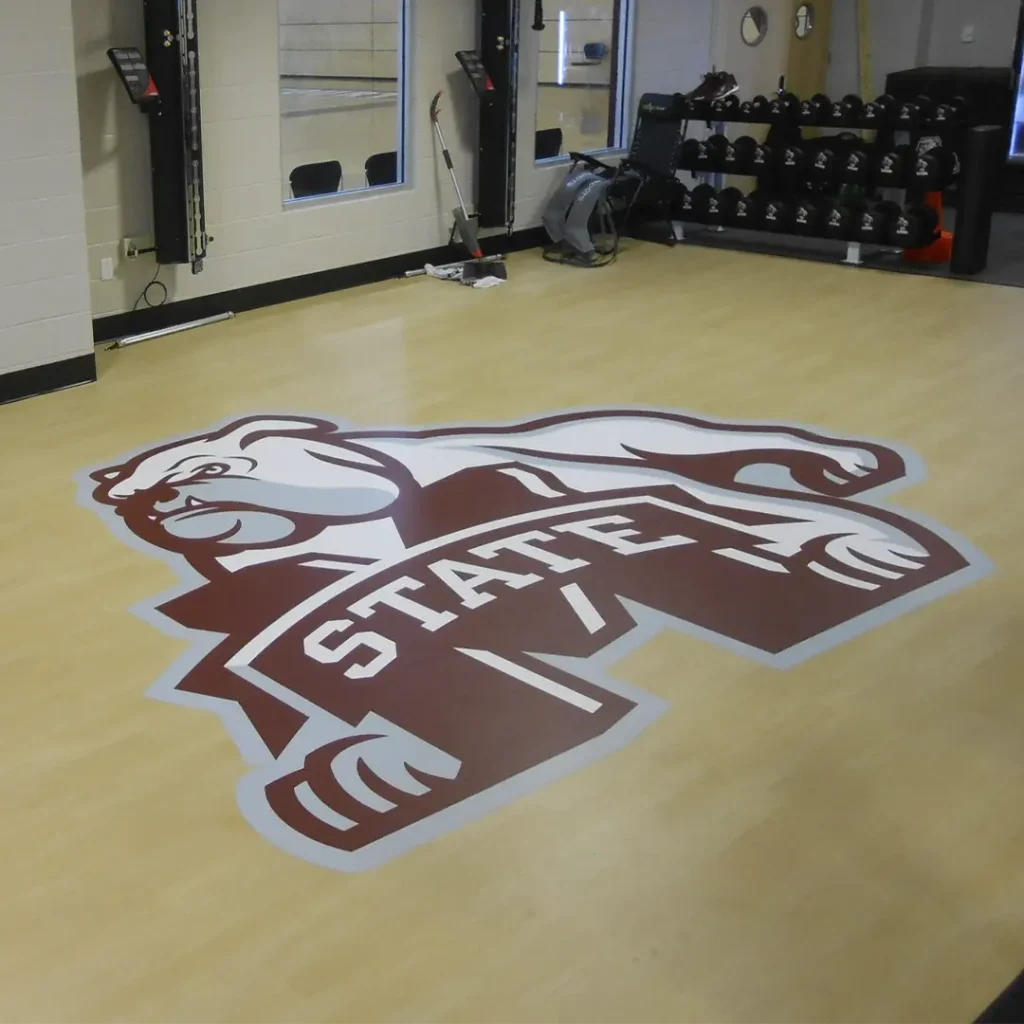 Bulldog engineered floor logo installed on wood-look vinyl flooring in a small fitness space with a dumbbell rack.