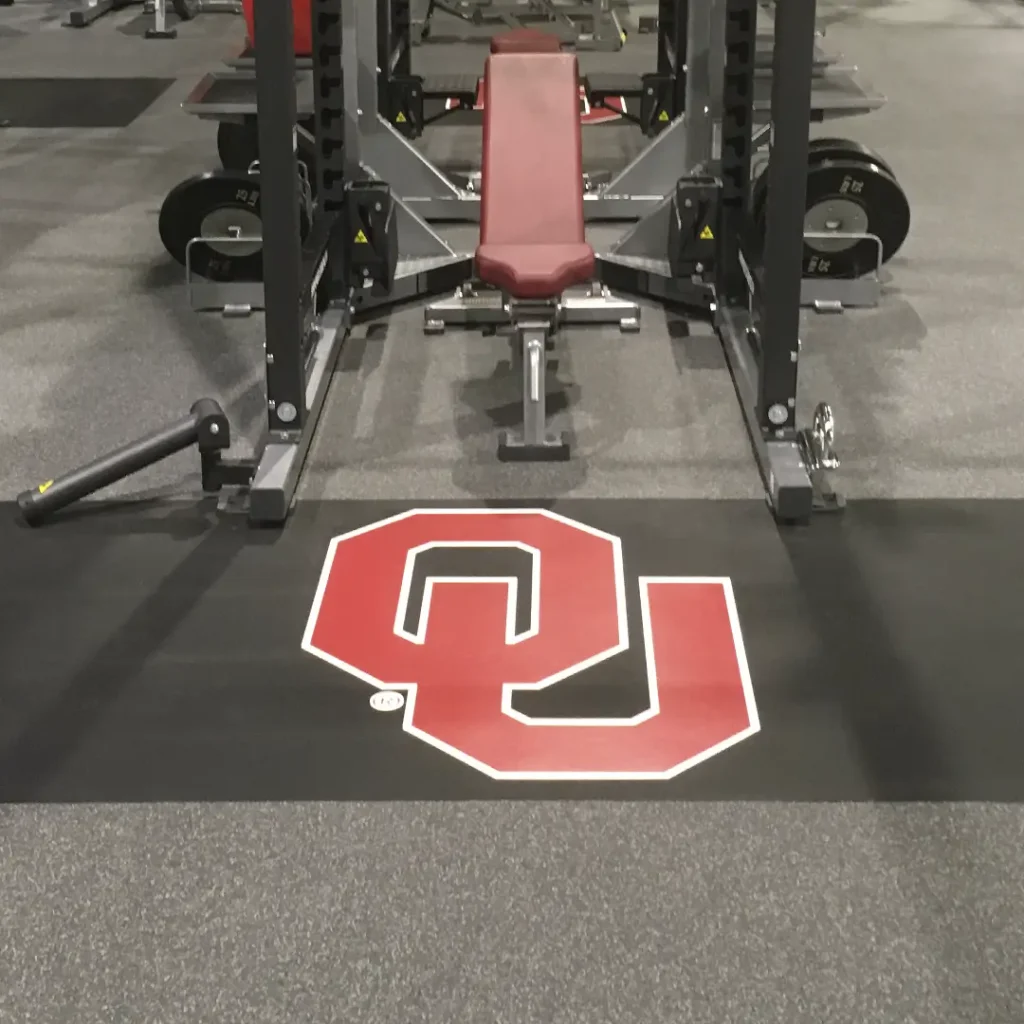 Inlaid OU monogram floor graphic installed on rubber flooring within a collegiate weight room platform