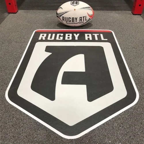 Close-up of a Rugby ATL engineered floor logo installed on rubber flooring, with a Rugby ATL ball placed above the graphic.